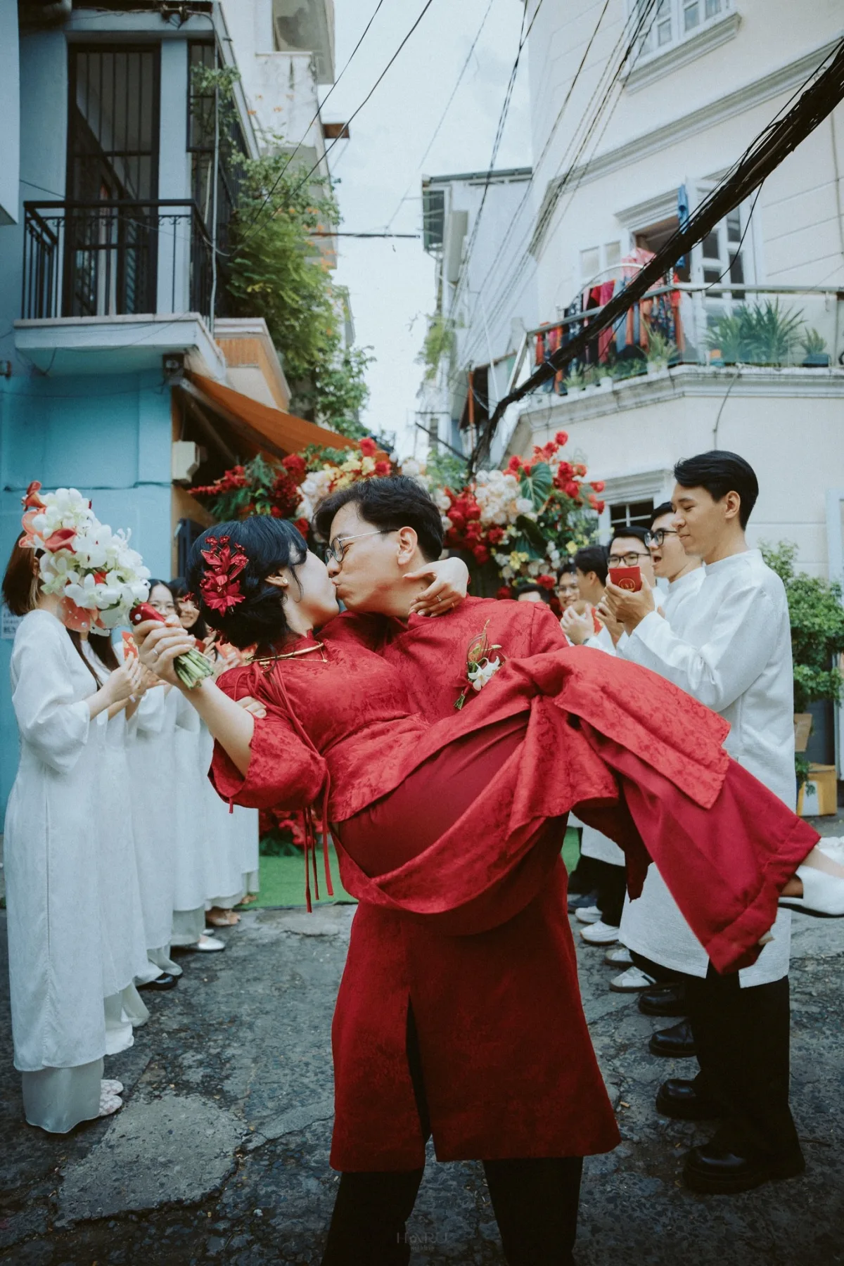 Chú rể bế cô dâu trong áo dài đỏ giữa gia đình và bạn bè